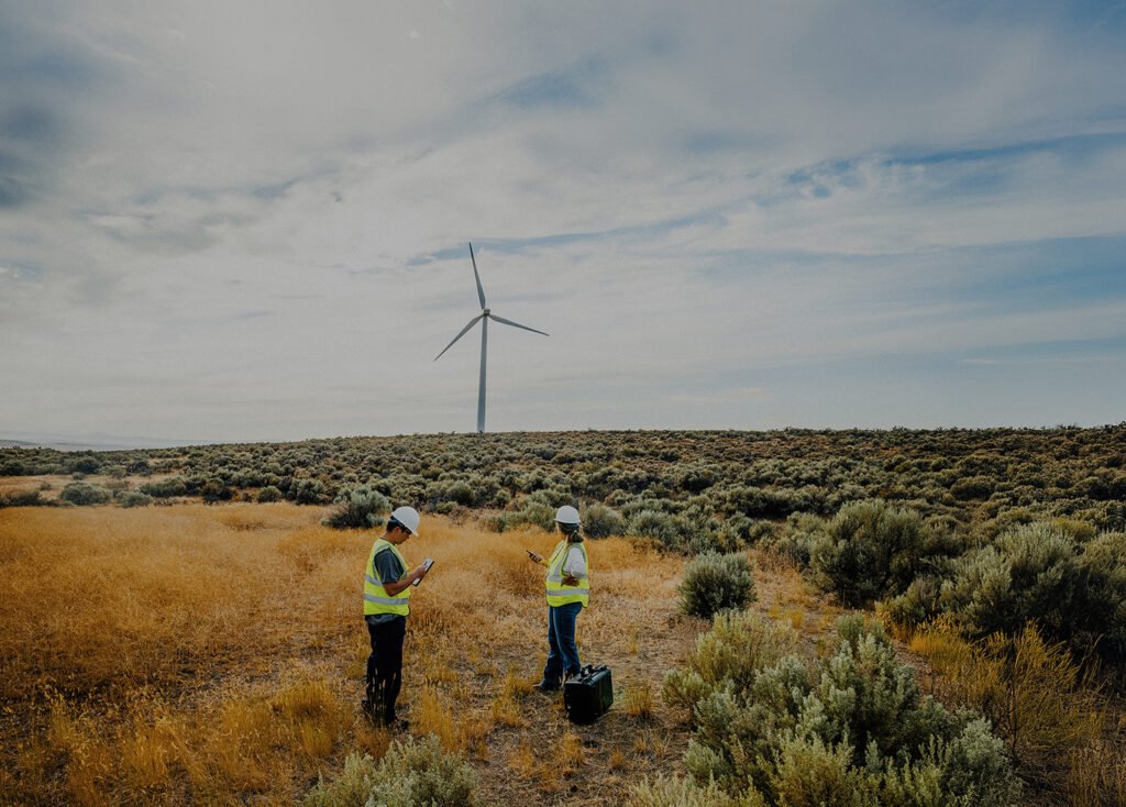 Two workers in safety vets and hard hats stand in a field.