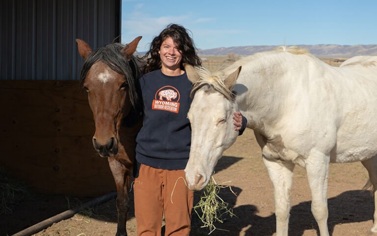 A woman stands, smiling, between two horses.
